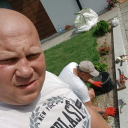 Prace ogrodnicze - Dwóch mężczyzn podczas układania obrzeży rabatki w ogrodzie. Jeden z nich robi selfie, drugi pracuje w tle. Widoczne narzędzia ogrodnicze, trawnik i fragment domu.