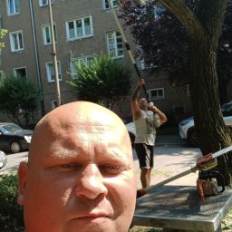 Prace ogrodnicze - Mężczyzna robiący selfie podczas pracy przycinania drzewa za pomocą piły na wysięgniku; w tle drugi mężczyzna z piłą spalinową i blok mieszkalny.