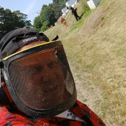 Prace ogrodnicze - Autoportret pracownika w ochronnej masce i kasku Kappa, ubrany w czerwoną koszulę w kratę, z widocznymi śladami trawy, w tle dwóch innych pracowników na zboczu trawnika.