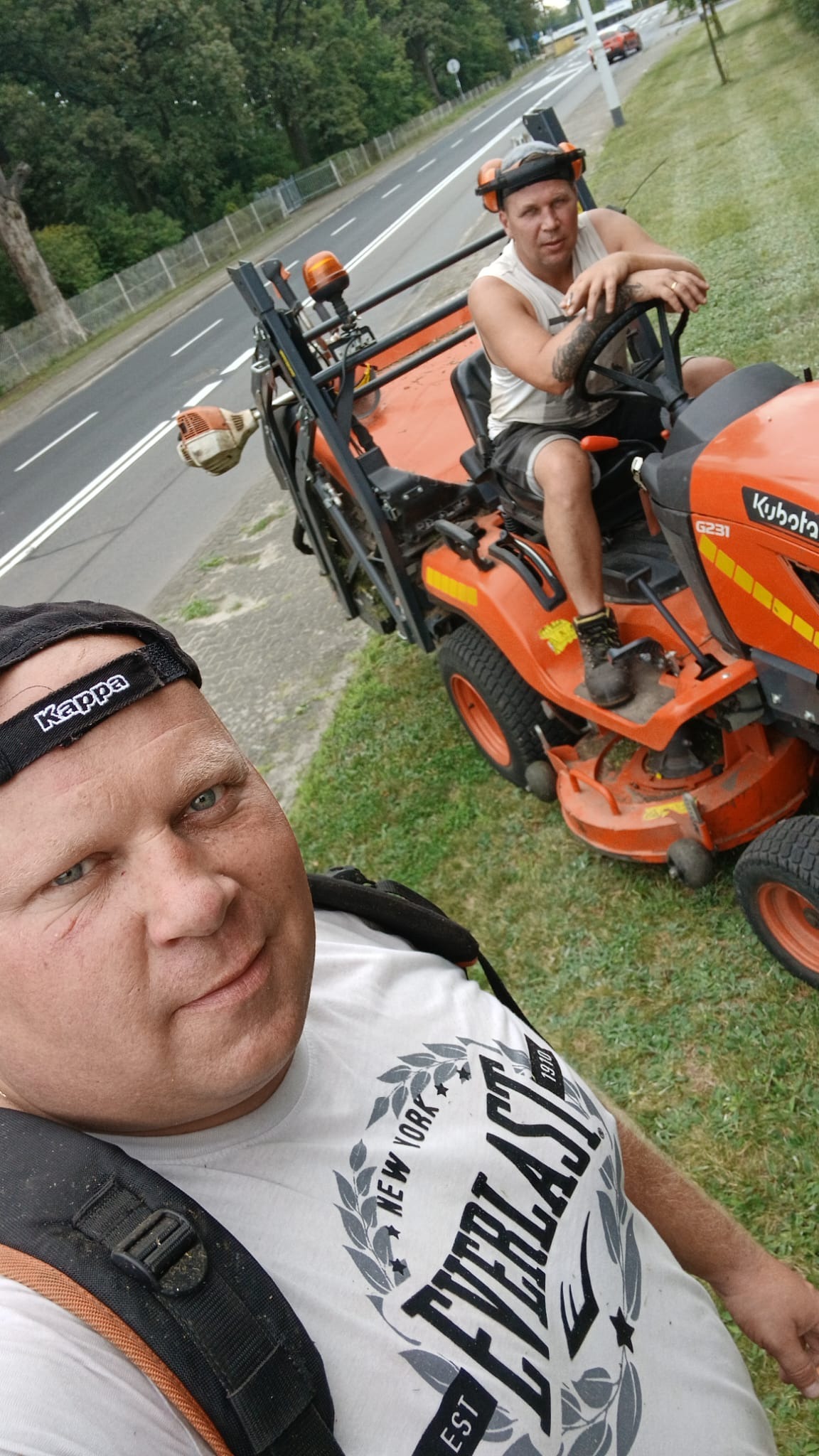 Mężczyzna robiący selfie na tle pomarańczowego traktora do trawy Kubota G231 z zamontowanym urządzeniem, drugi mężczyzna siedzi na traktorze, w tle droga i drzewa.