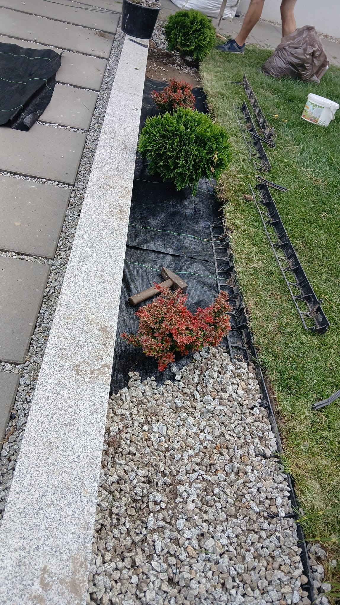 Fragment ogrodu w trakcie tworzenia: obrzeża z kostki granitowej, czarna agrowłóknina, ozdobne krzewy, wysypany żwir, plastikowe obrzeża trawnikowe i młotek.
