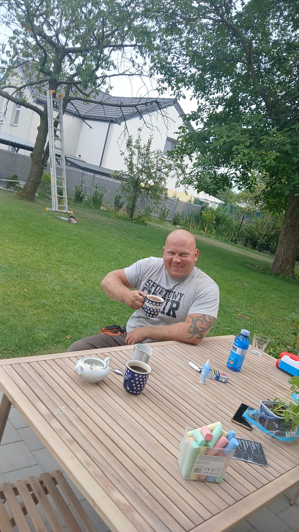 Uśmiechnięty mężczyzna z tatuażami, w roboczym ubraniu, siedzi przy drewnianym stole w ogrodzie, trzymając filiżankę. W tle trawnik, drzewa i drabina oparta o drzewo.