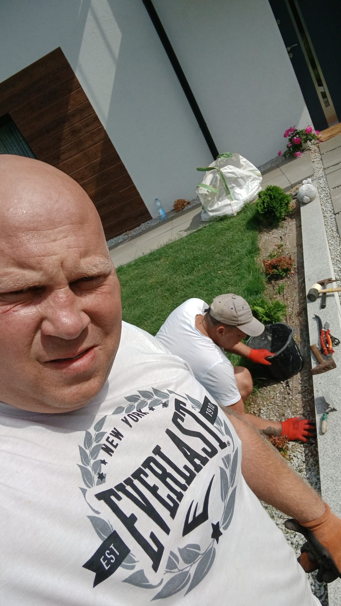 Dwóch mężczyzn podczas układania obrzeży rabatki w ogrodzie. Jeden z nich robi selfie, drugi pracuje w tle. Widoczne narzędzia ogrodnicze, trawnik i fragment domu.