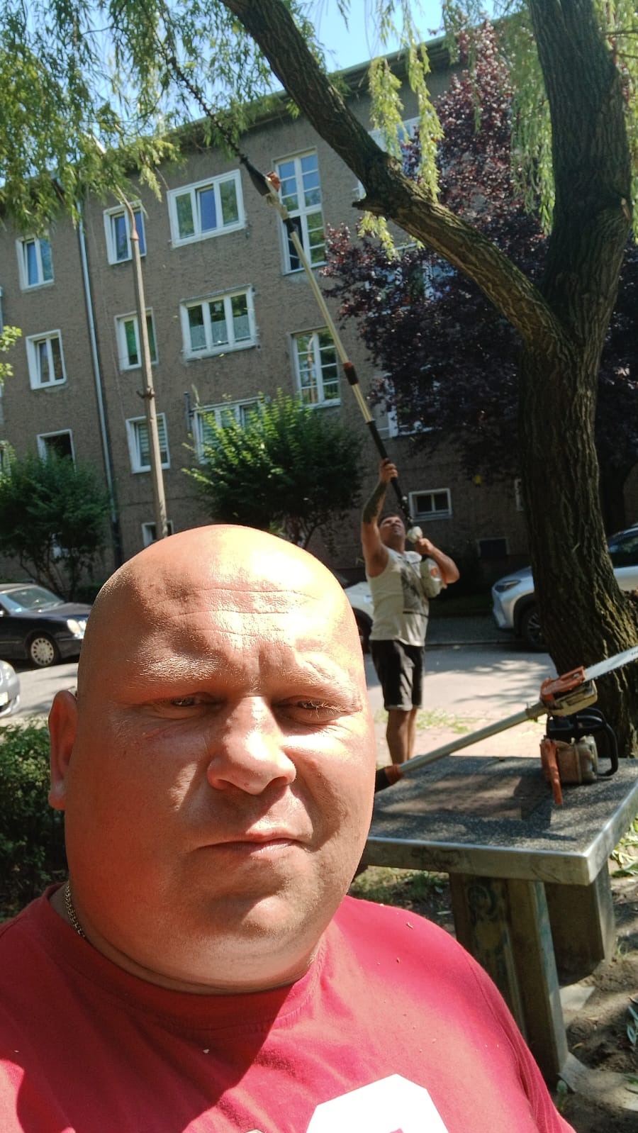 Mężczyzna robiący selfie podczas pracy przycinania drzewa za pomocą piły na wysięgniku; w tle drugi mężczyzna z piłą spalinową i blok mieszkalny.