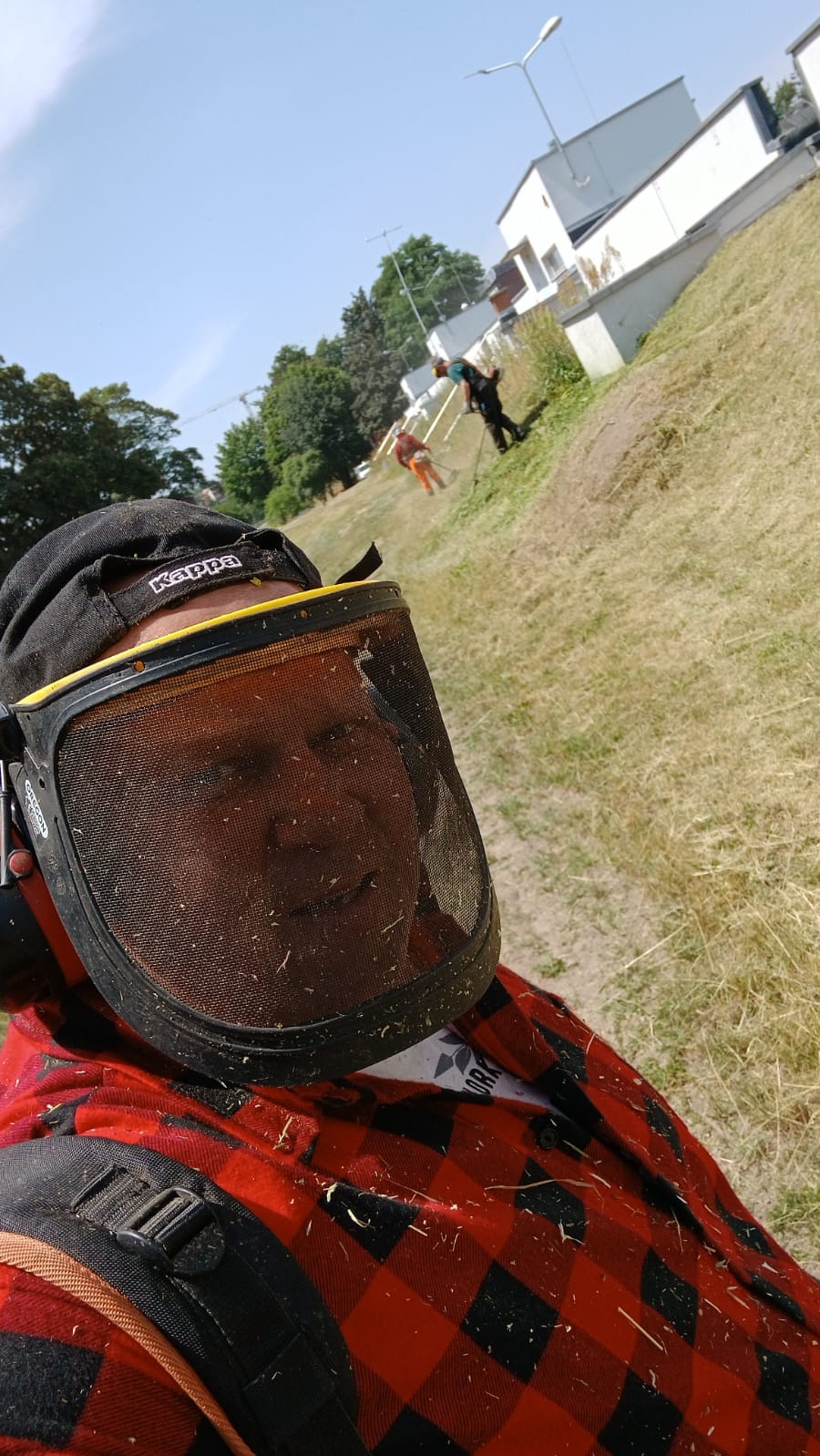 Autoportret pracownika w ochronnej masce i kasku Kappa, ubrany w czerwoną koszulę w kratę, z widocznymi śladami trawy, w tle dwóch innych pracowników na zboczu trawnika.