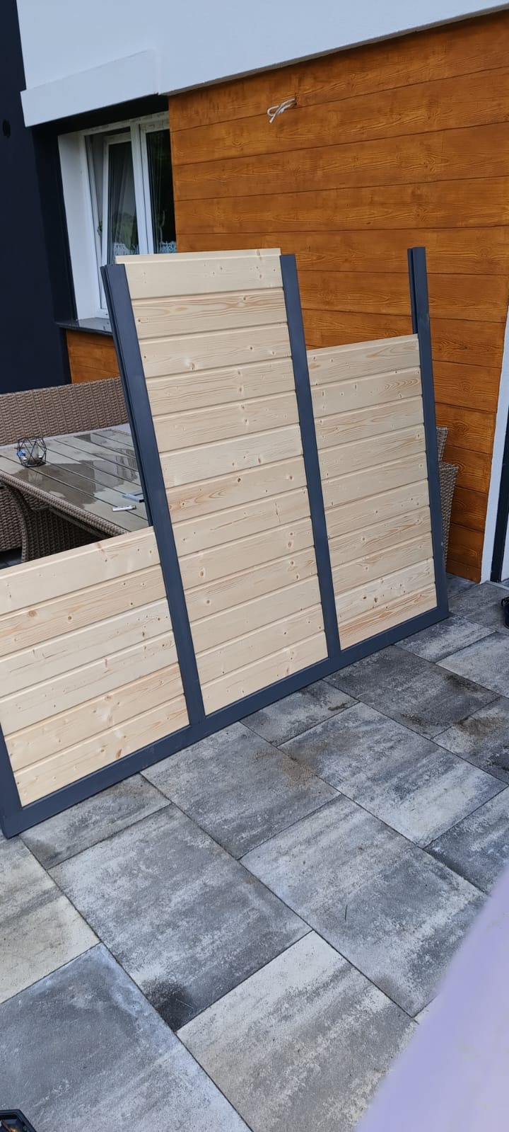 Drewniane panele ogrodzeniowe z szarą ramą, ustawione na szarej kostce brukowej, w tle brązowa drewniana ściana budynku i fragment rattanowego kompletu mebli.