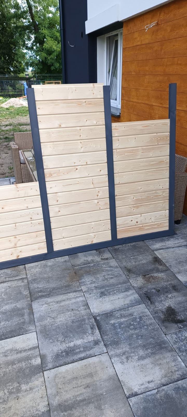 Drewniany parawan ogrodowy z szarą ramą, ustawiony na szarym bruku, na tle domu z drewnianą elewacją.