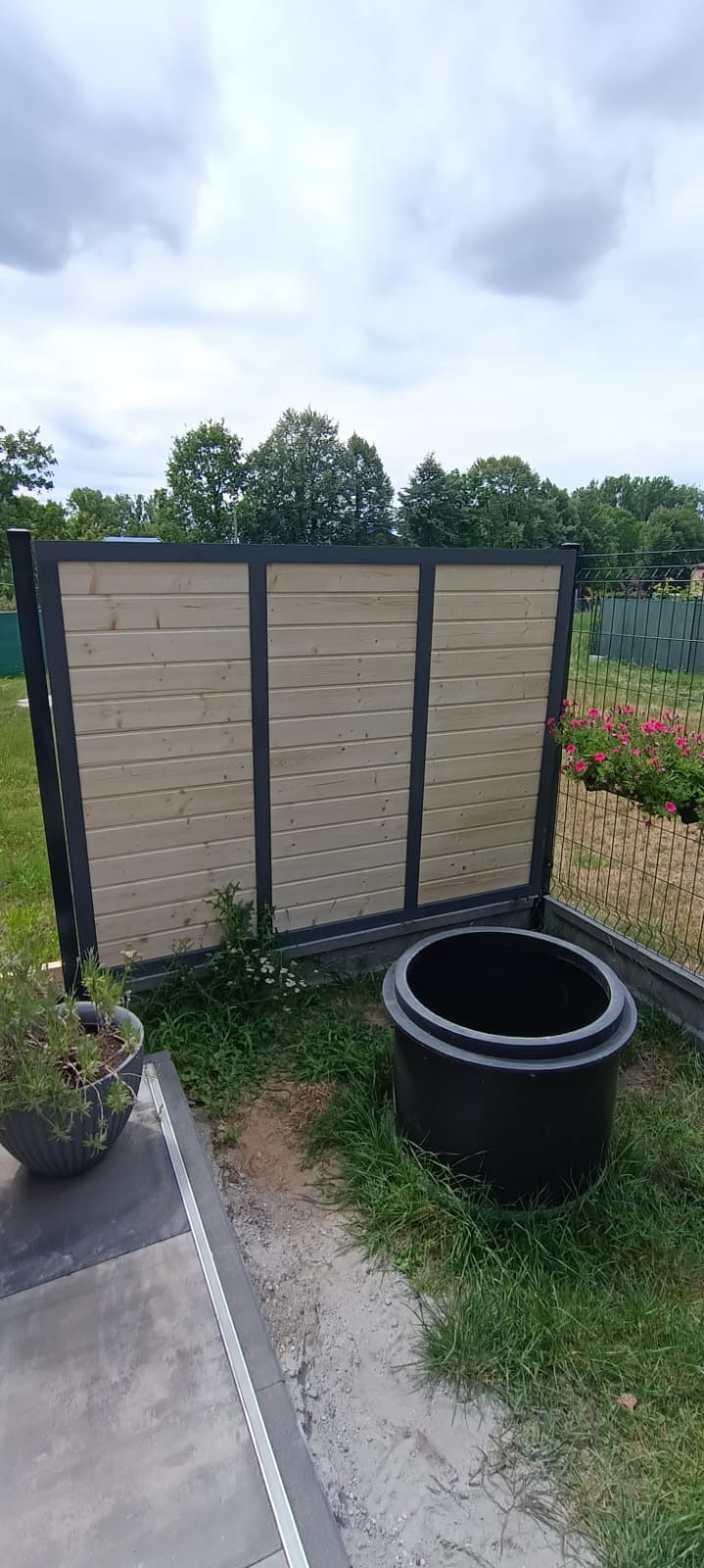 Nowoczesny element ogrodowy: drewniana przesłona z metalową ramą, obok czarny zbiornik retencyjny, donica z ziołami i fragment betonowej ścieżki w ogrodzie.