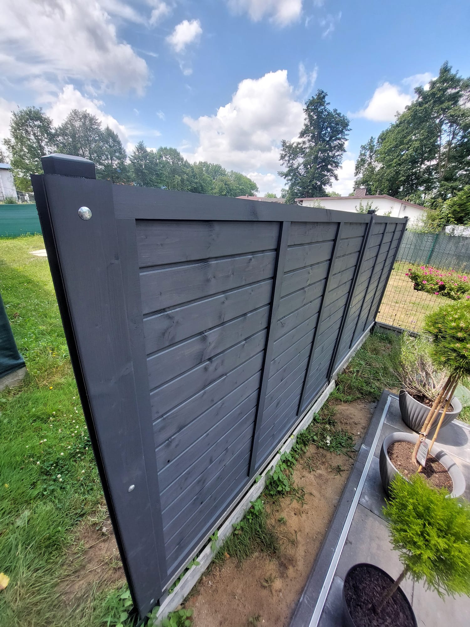 Nowoczesny, pomalowany na grafitowo drewniany płot panelowy, oddzielający trawnik od rabaty z roślinami w ogrodzie, na tle błękitnego nieba z chmurami.