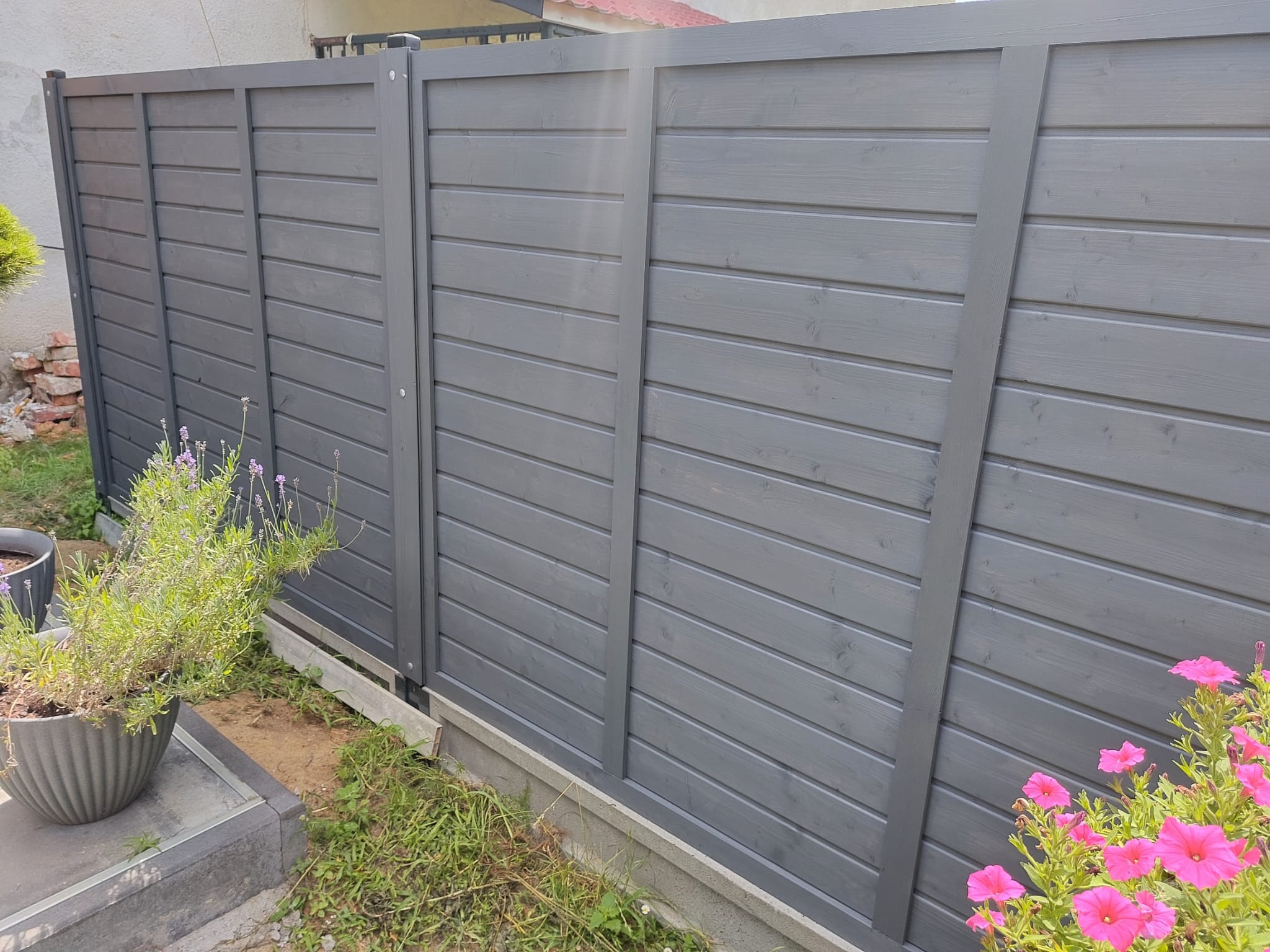 Nowoczesne, szare ogrodzenie panelowe z drewna, z widocznymi kwiatami w donicach i na rabacie, częściowo na tle zieleni.