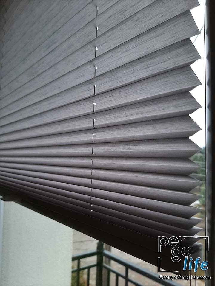 Szare, plisowane osłony okienne z widocznym logotypem producenta w prawym dolnym rogu, zamontowane w oknie z widokiem na balkon.