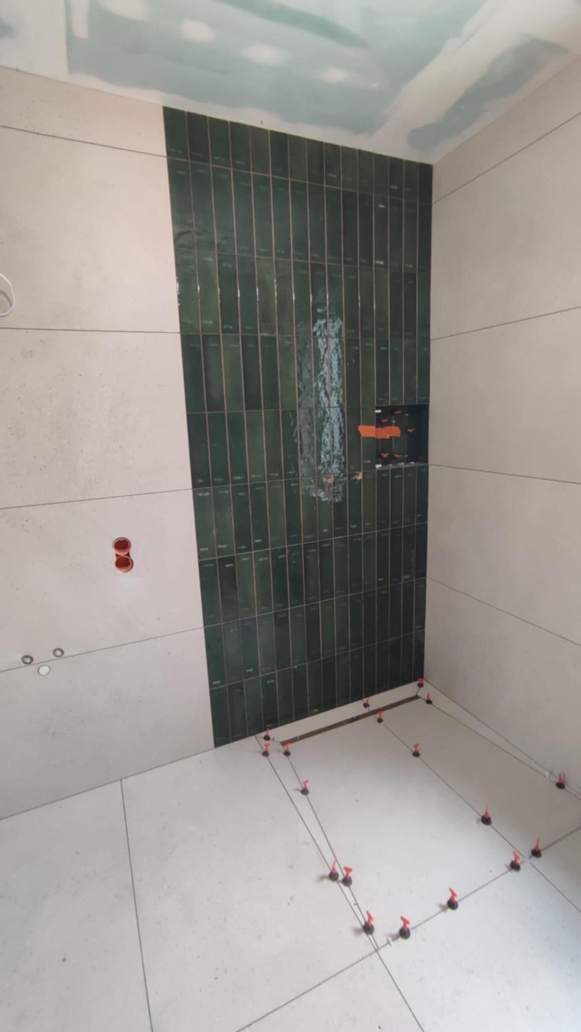 Wnętrze łazienki w trakcie prac glazurniczych: ułożone zielone, pionowe płytki na ścianie, poziomowanie płytek podłogowych z użyciem klipsów i klinów. Widoczne puszki elektryczne.