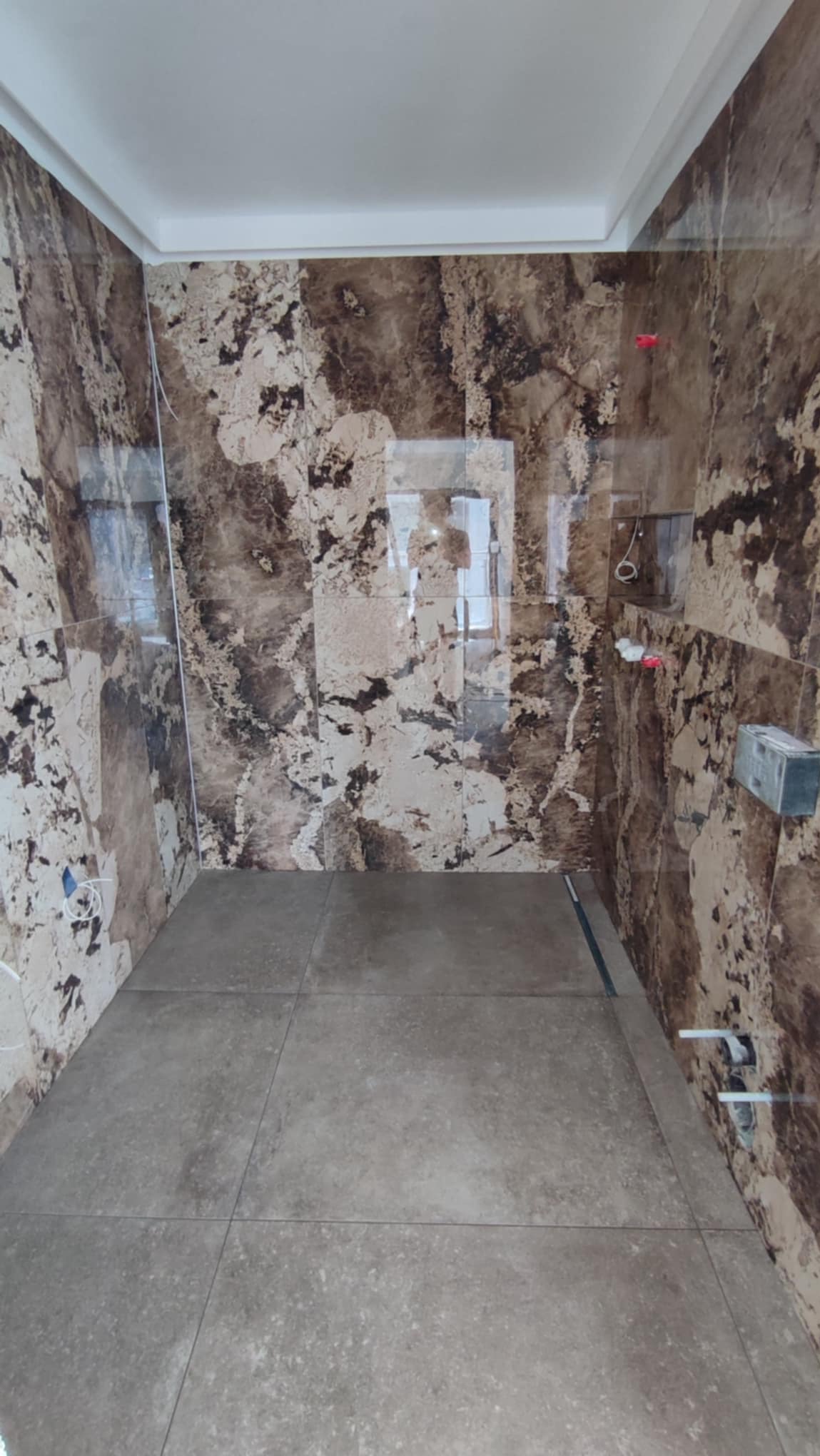 Wykończona łazienka z brązowymi płytkami imitującymi marmur na ścianach i szarą podłogą z płytek. Widoczne elementy instalacji wodnej.