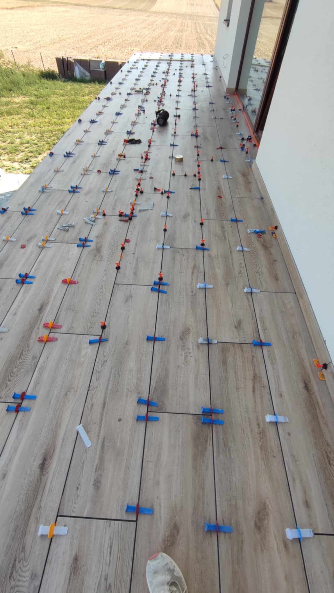 Układanie płytek imitujących drewno na tarasie, z użyciem systemu poziomowania. Widoczne krzyżyki dystansowe i kliny. W tle trawa i budynek.