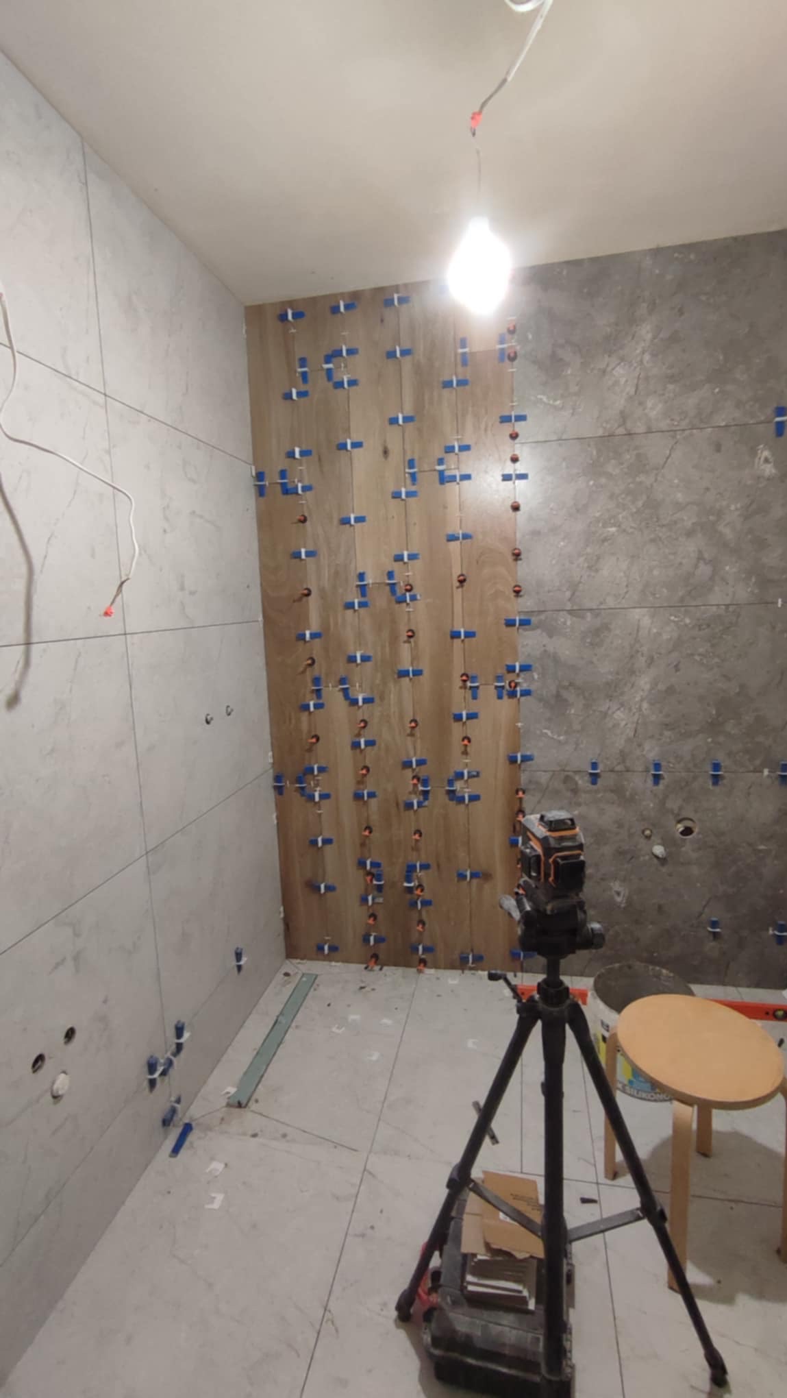Łazienka w trakcie wykańczania: częściowo ułożone płytki imitujące drewno, poziomica laserowa na statywie, krzyżyki dystansowe, widoczne instalacje.