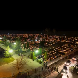 Nocny widok na cmentarz oświetlony tysiącami zapalonych zniczy, widoczne alejki, drzewa i zaparkowane samochody, wysoka perspektywa.