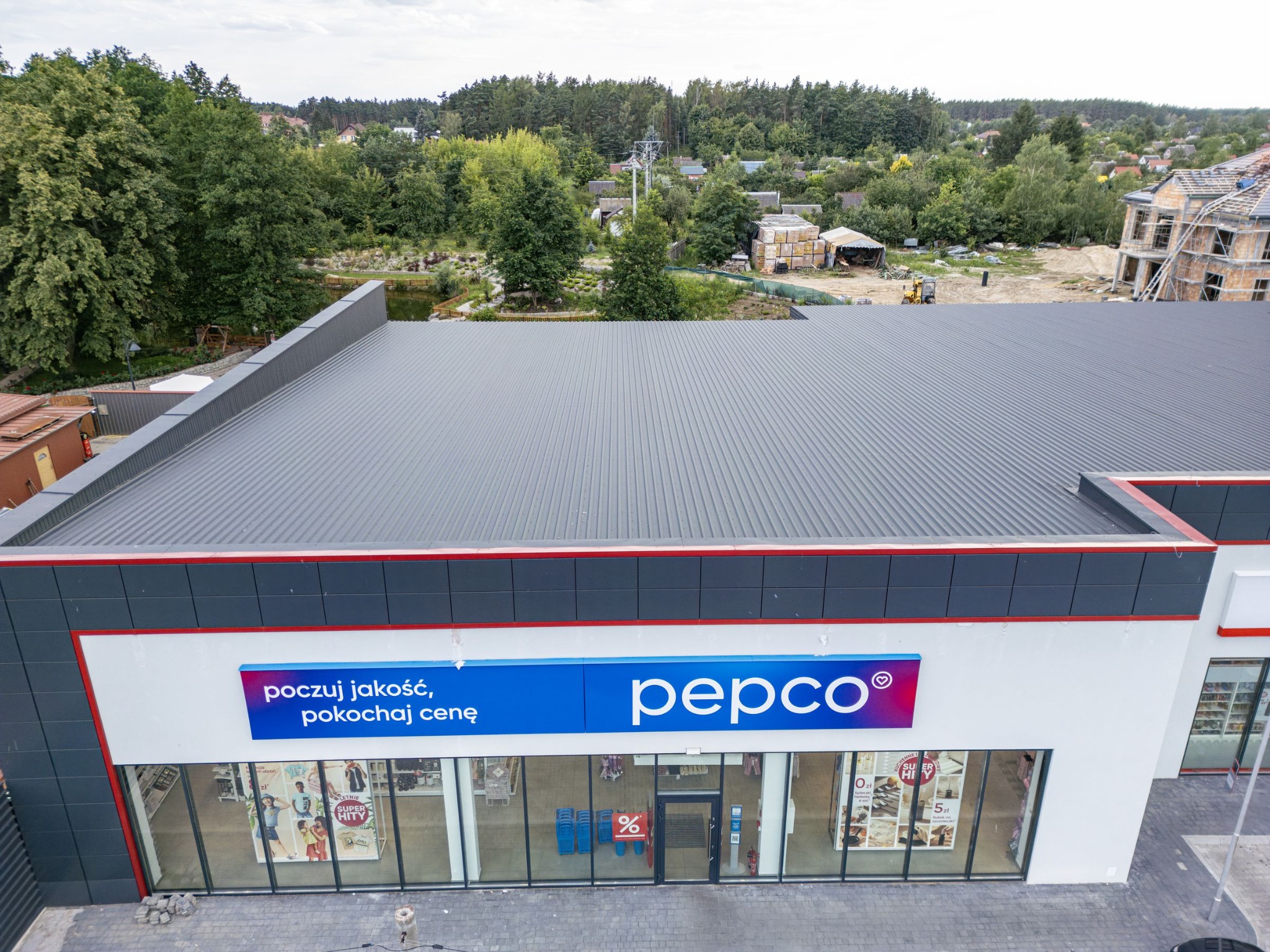 Zdjęcie z drona nowo otwartego sklepu Pepco z szarym dachem na tle zieleni i budowy, widoczne logo i wnętrze sklepu przez duże okna wystawowe.