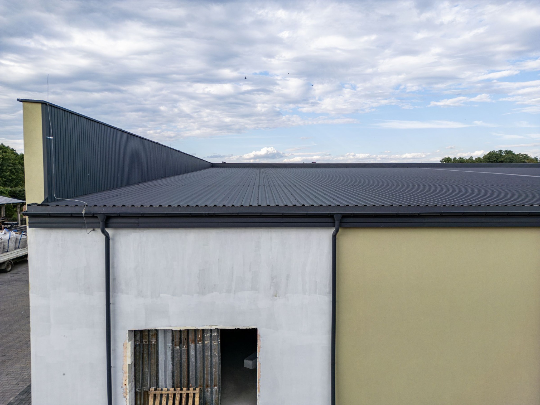 Budynek magazynowy z szarą blachodachówką trapezową, widoczne orynnowanie, ściany otynkowane na szaro i beżowo, częściowo zakryte okno zabezpieczone metalowymi prętami, na zewnątrz zaparkowany...