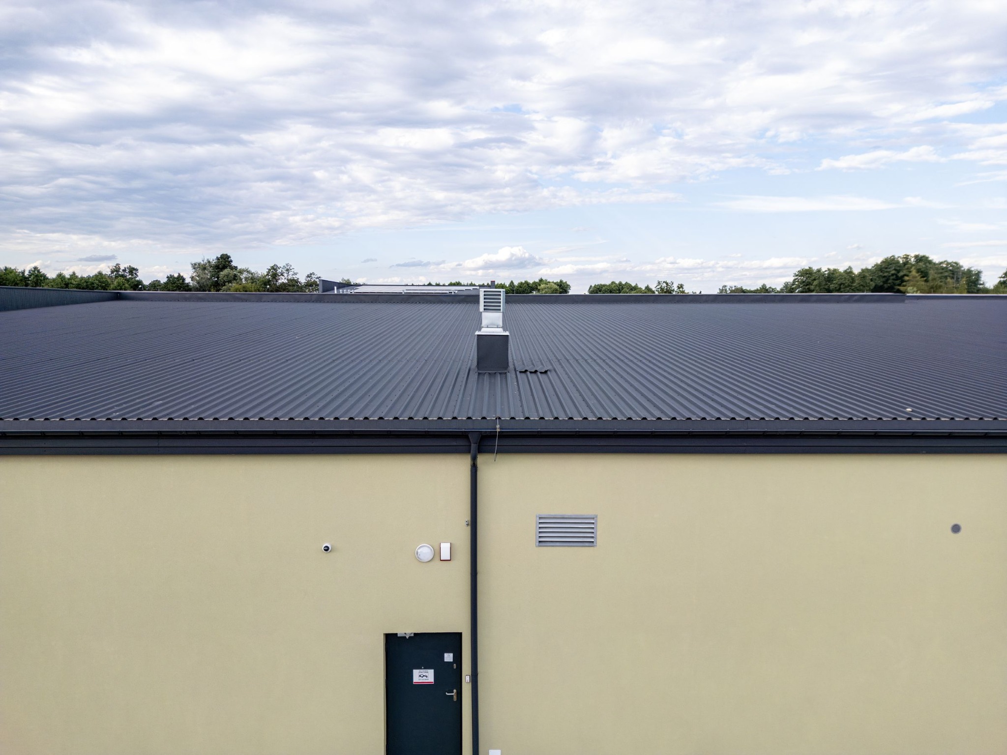 Szary dach z blachy trapezowej z kominem wentylacyjnym na budynku o jasnej elewacji, widoczne rynny, drzwi wejściowe i kratka wentylacyjna.