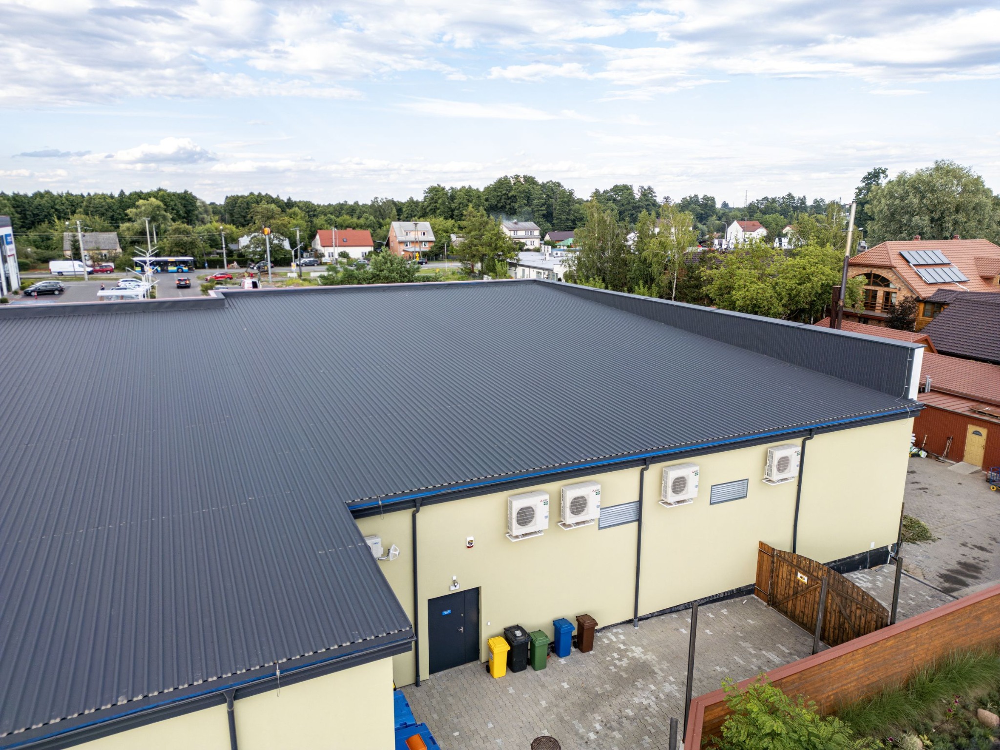 Widok z góry na budynek z szarą blachodachówką, żółtą elewacją, klimatyzatorami na ścianie i rzędem kolorowych pojemników na śmieci przy wejściu. W tle zabudowa jednorodzinna i drzewa.