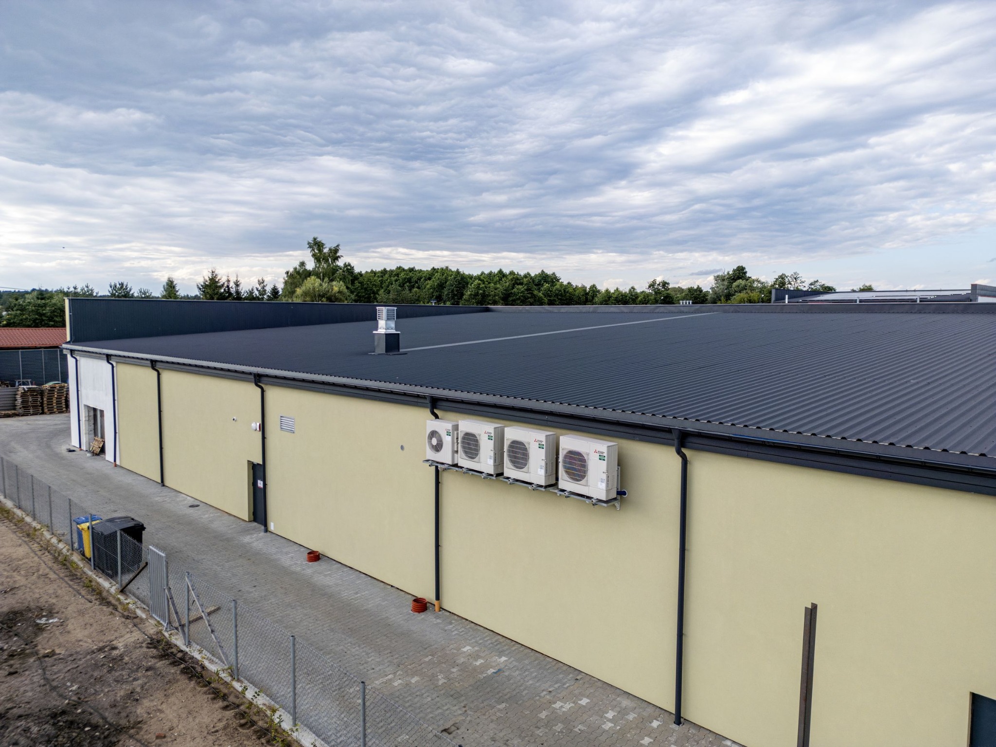 Widok z góry na nowo wybudowany magazyn z beżową elewacją, szarą blachodachówką, czterema klimatyzatorami na ścianie i ogrodzeniem z siatki.