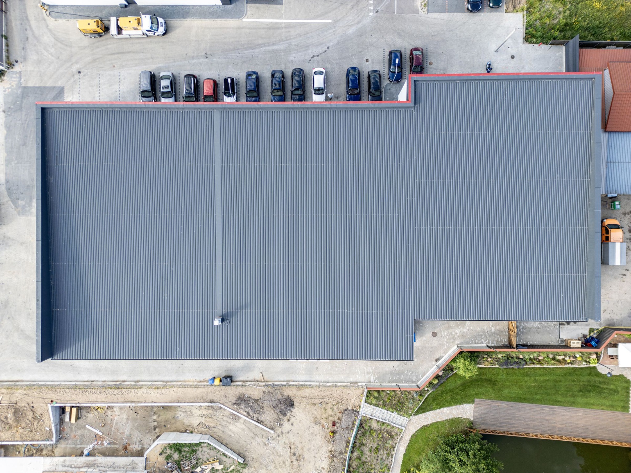 Zdjęcie z lotu ptaka przedstawia duży budynek przemysłowy z szarym, blaszany dachem trapezowym, zaparkowane samochody wzdłuż dachu, teren budowy i zagospodarowany teren zielony z oczkiem wodnym.