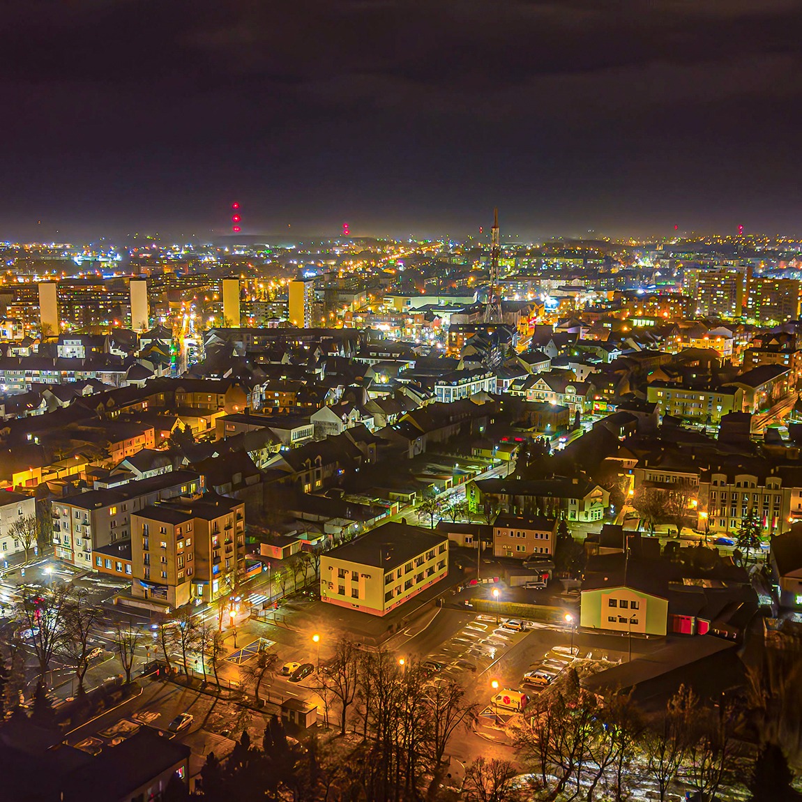Nocna panorama miasta z widokiem na oświetlone budynki, ulice i parking, sfotografowana z góry.