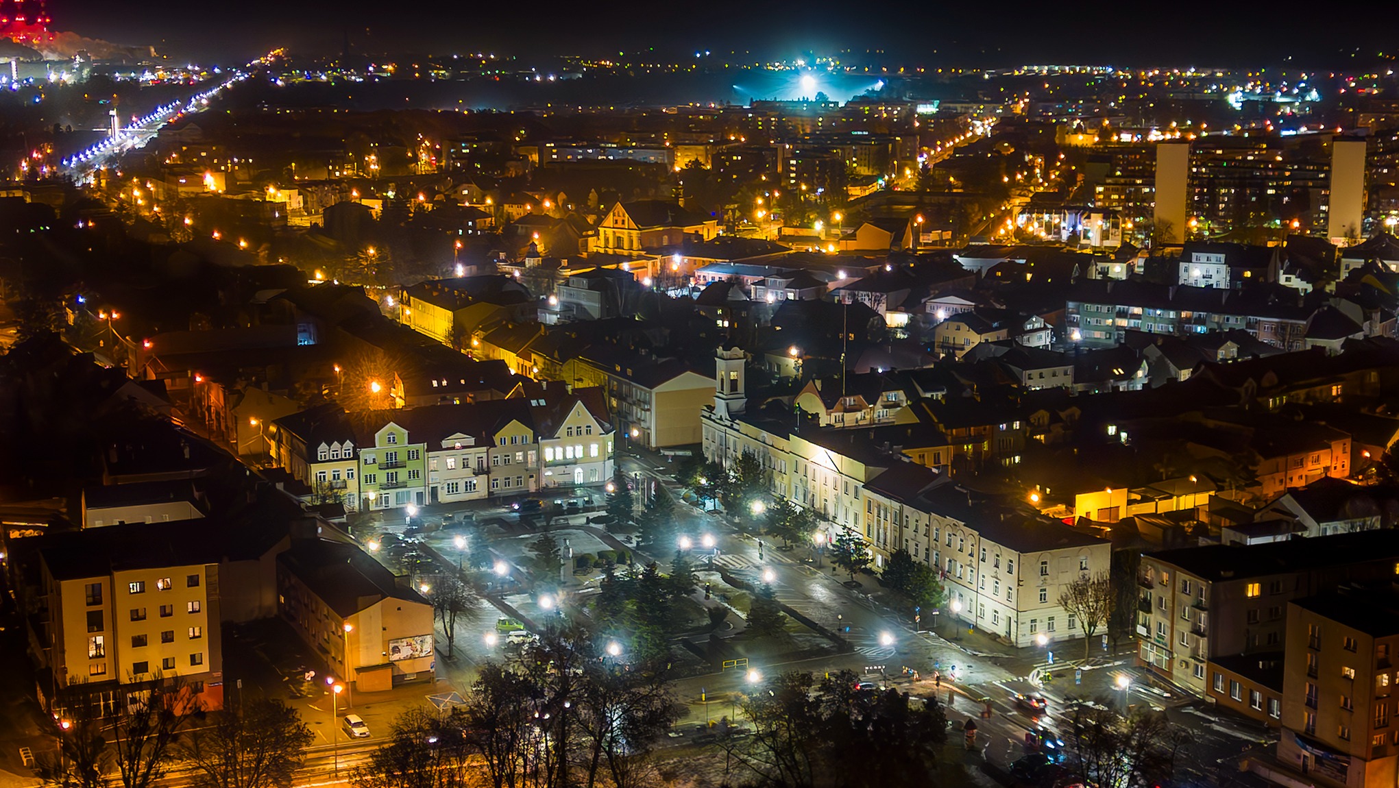 Nocna panorama miasta z lotu ptaka, ukazująca oświetlone budynki, ulice i plac, z widocznymi latarniami i samochodami w ruchu.