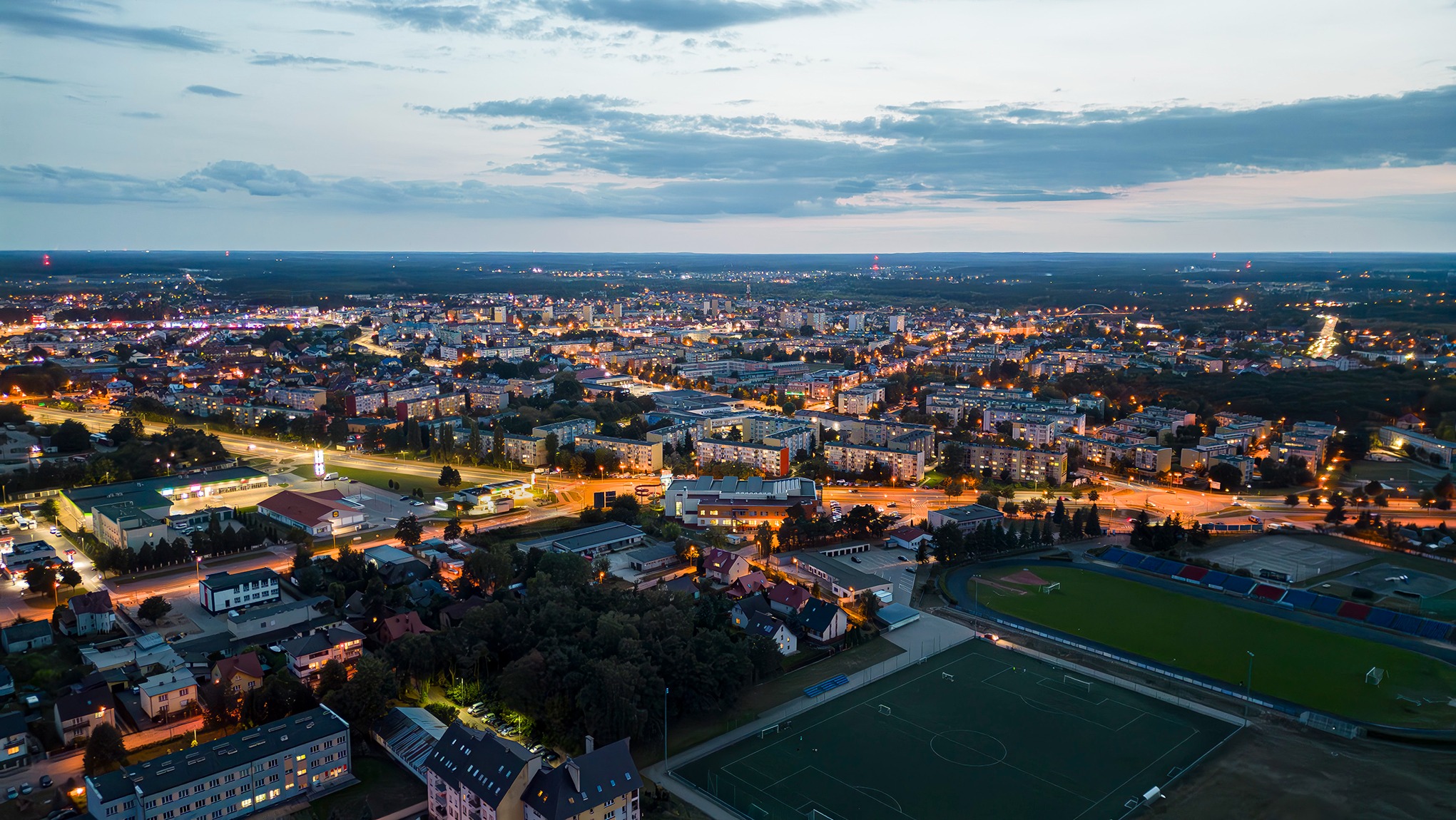 Panorama miasta z lotu ptaka o zmierzchu, widoczne oświetlone budynki mieszkalne, stadion i drogi.