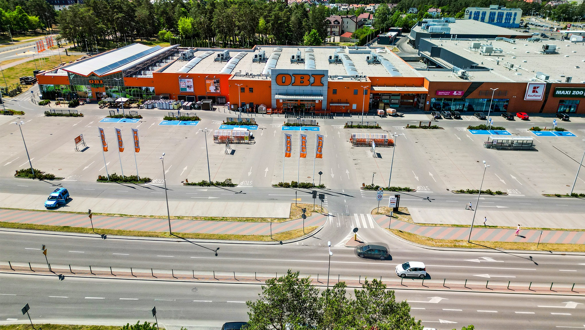 Zdjęcie z lotu ptaka przedstawia budynek OBI z dużym parkingiem, flagami reklamowymi i pobliską drogą z samochodami, otoczone zielenią drzew i zabudową mieszkalną.