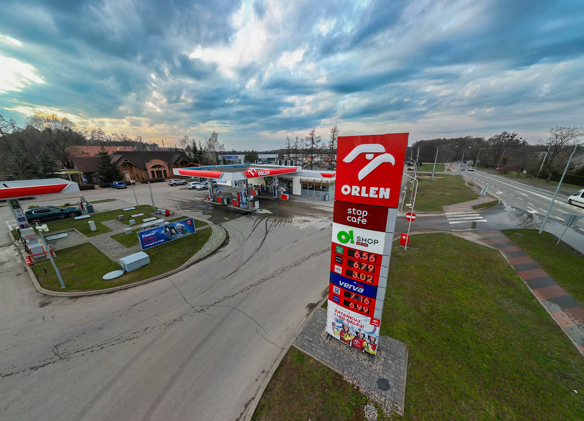 Stacja benzynowa Orlen z dystrybutorami, budynkiem sklepu i pylonem cenowym na tle pochmurnego nieba, widok z góry.