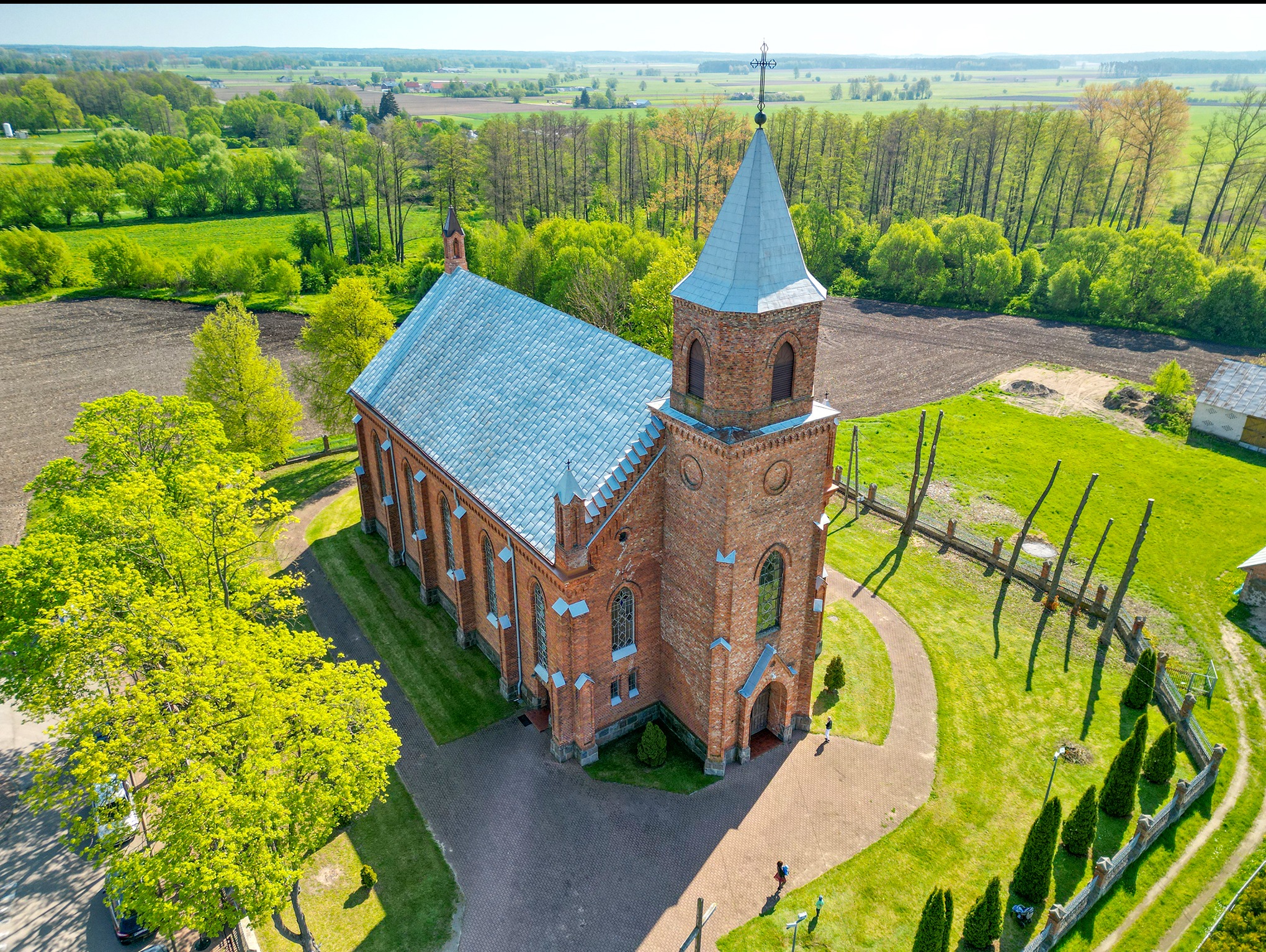 Zdjęcie z lotu ptaka przedstawia ceglany kościół z szarym dachem i wieżą zwieńczoną krzyżem, otoczony zielenią drzew i polami uprawnymi, z widocznym ogrodzeniem z drewnianych pali.