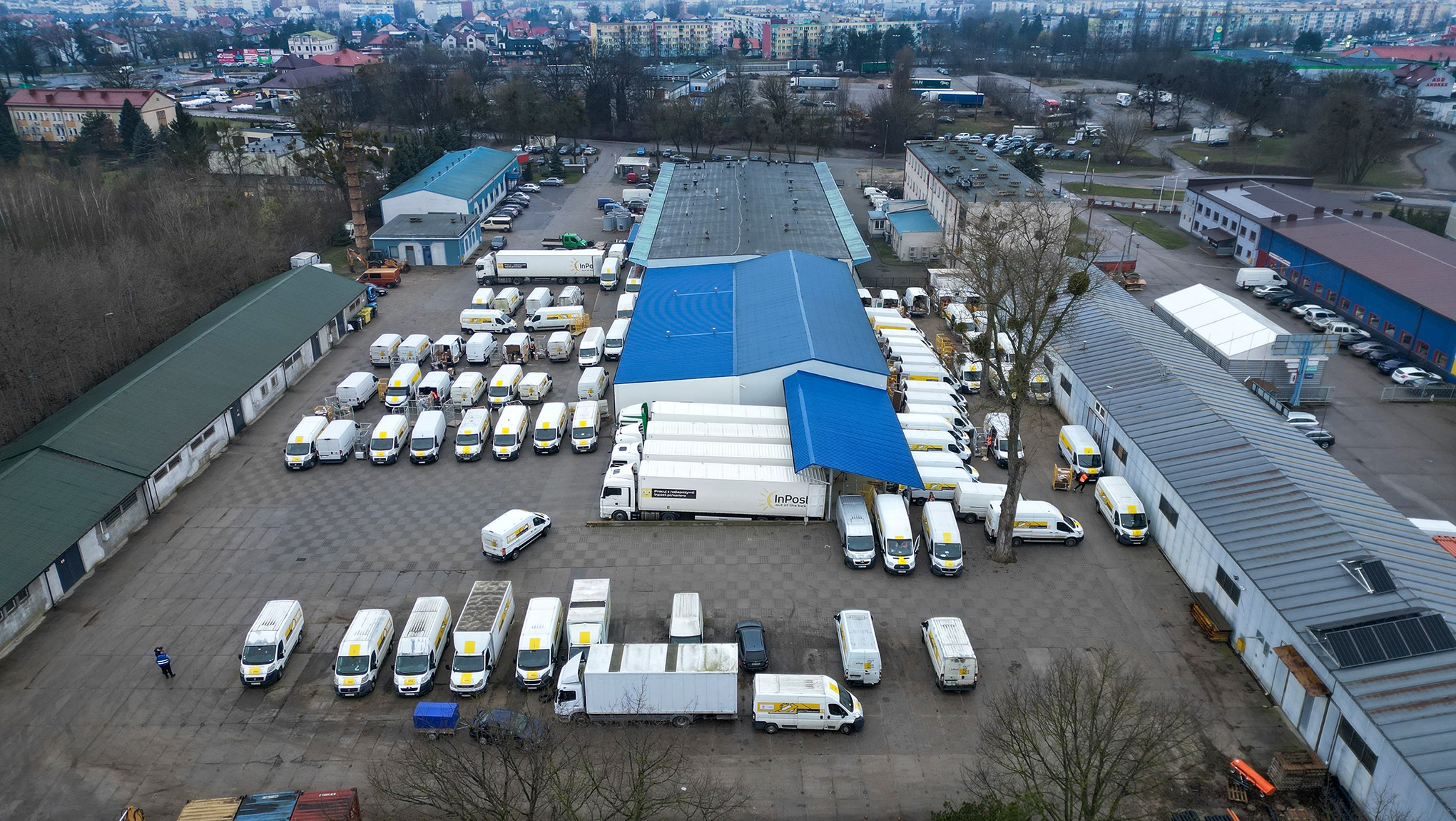 Widok z góry na duży parking z zaparkowanymi samochodami dostawczymi InPost, niebiesko-biały magazyn w tle, pracownik w uniformie na lewo od samochodów.