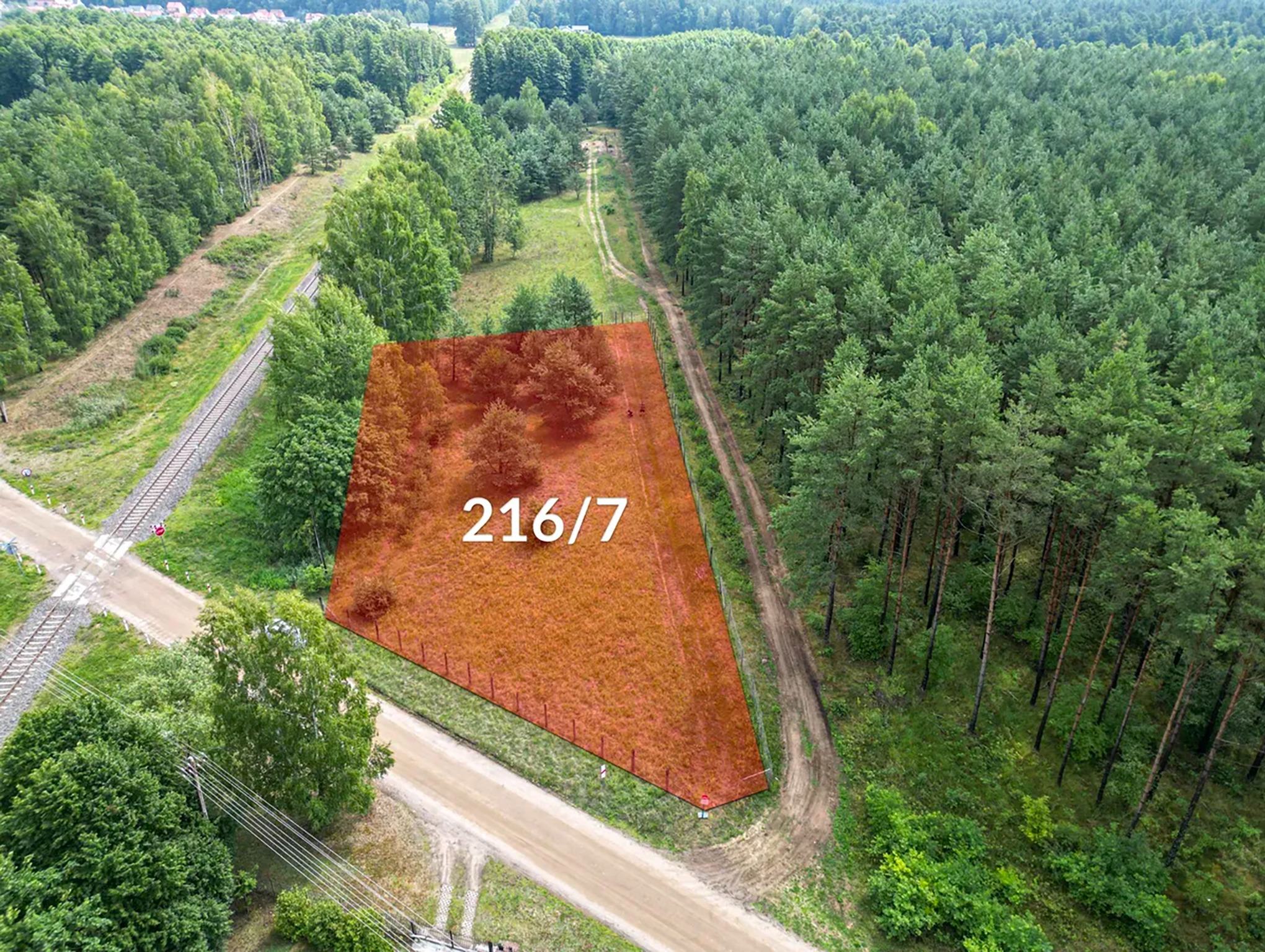 Zdjęcie z drona działki nr 216/7, otoczonej lasem i z linią kolejową w pobliżu, zaznaczonej czerwoną obwódką.
