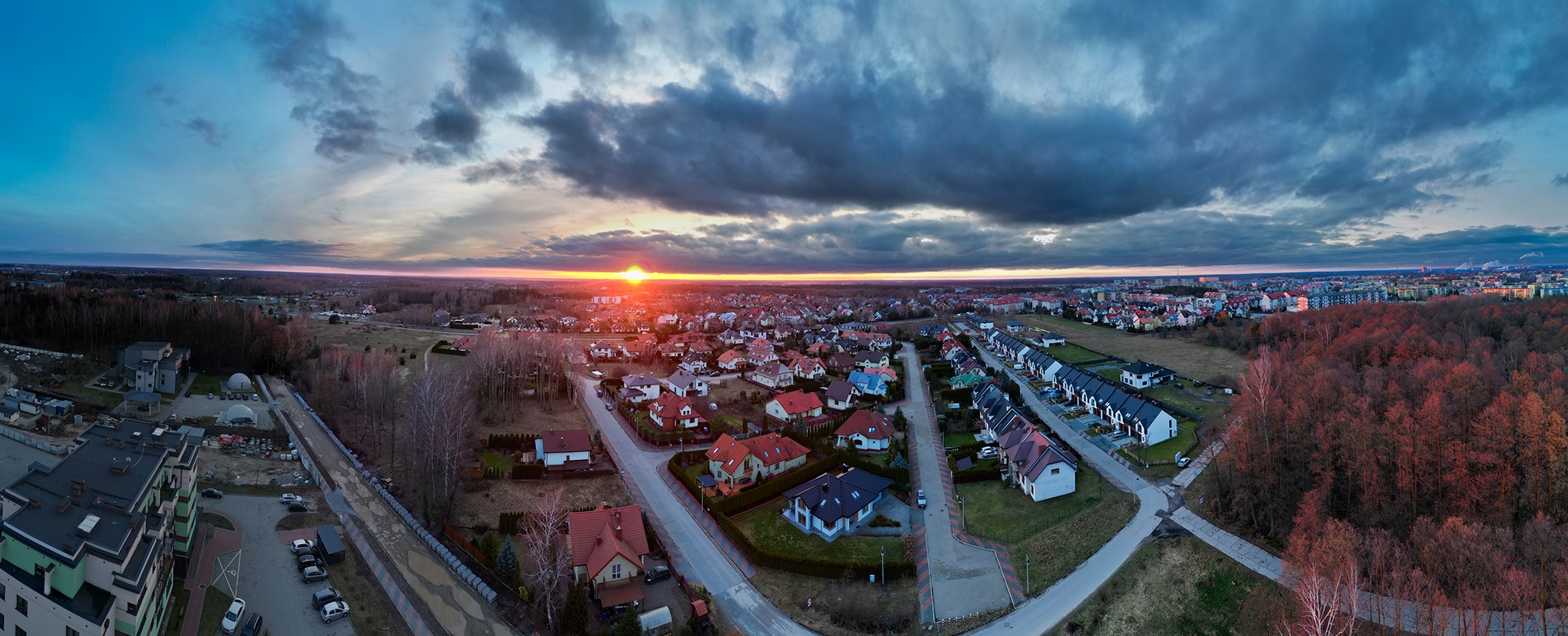 Panoramiczny widok z drona na osiedle domów jednorodzinnych i miasto o zachodzie słońca, z widocznymi ciemnymi chmurami, lasem w jesiennej kolorystyce i budynkami w oddali.