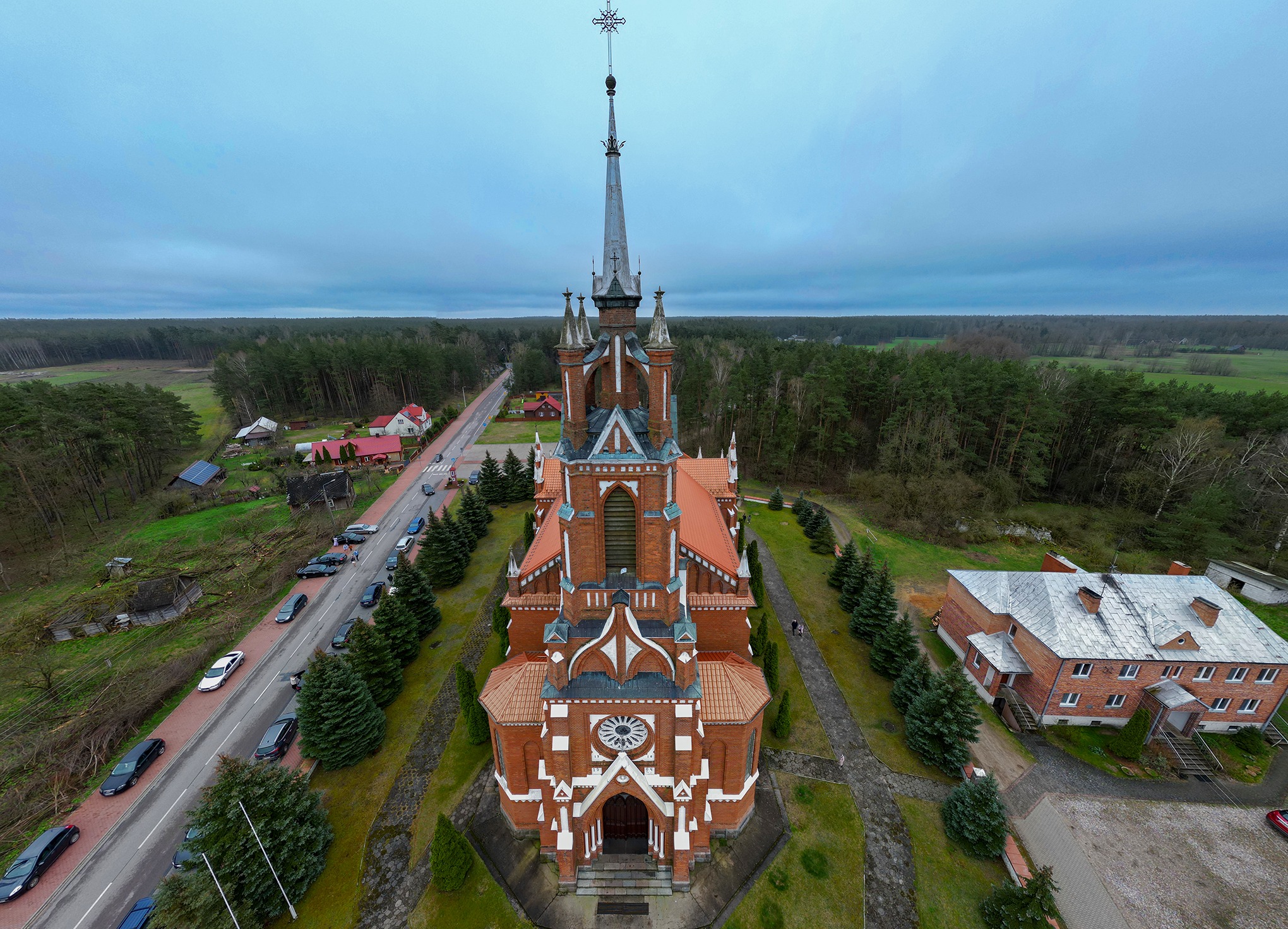 Widok z drona na ceglany kościół z wysoką wieżą i krzyżem na szczycie, otoczony zielenią i lasem, z drogą i samochodami w pobliżu.