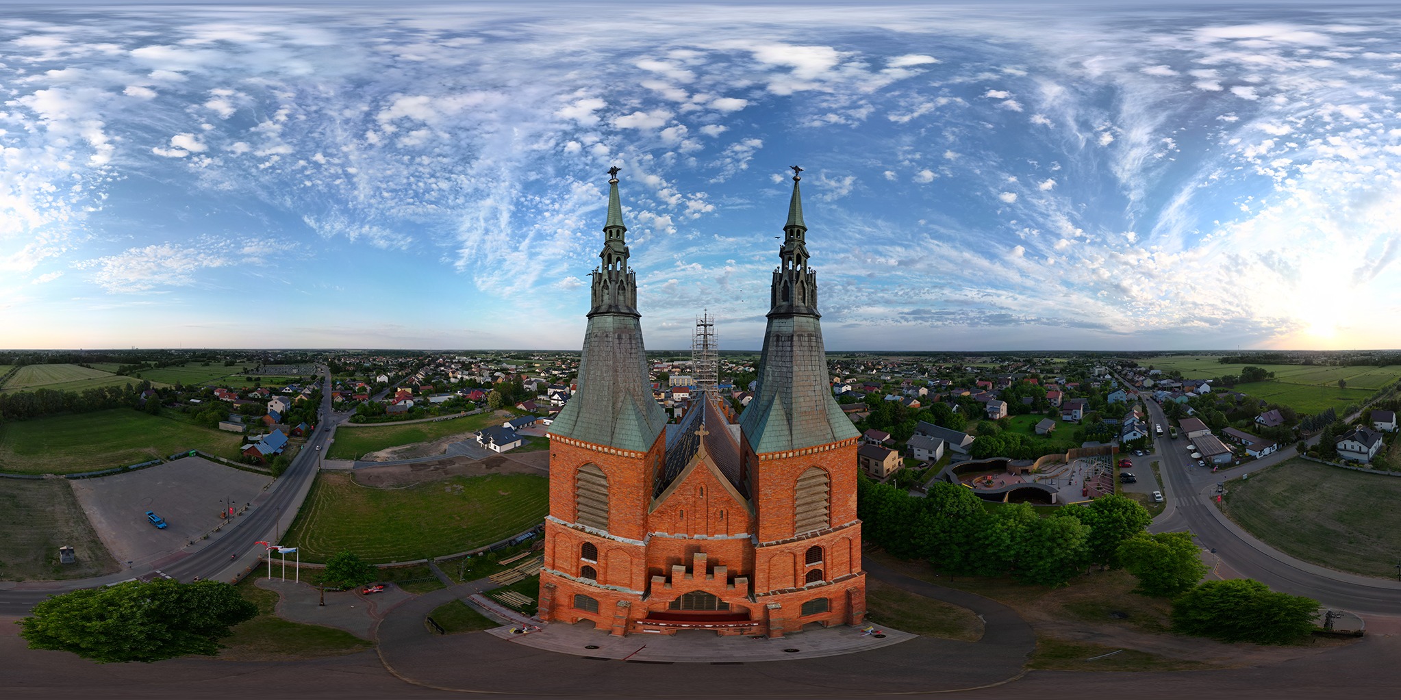 Panoramiczny widok z drona na ceglany kościół z dwiema wieżami, otoczony zielenią i zabudową miejską, pod błękitnym niebem z chmurami.