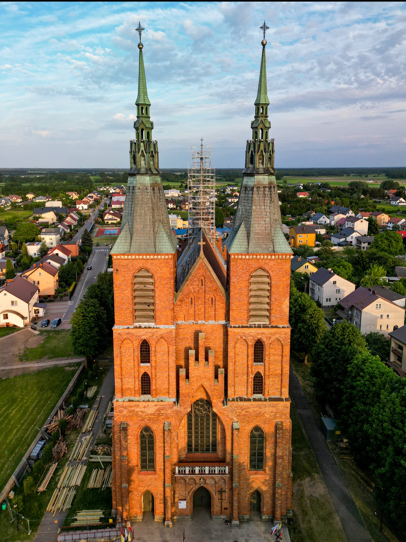 Zdjęcie z drona przedstawia ceglany kościół z dwiema wieżami zakończonymi zielonymi iglicami, w tle zabudowa mieszkalna i zieleń, na jednej z wież widoczne rusztowania.