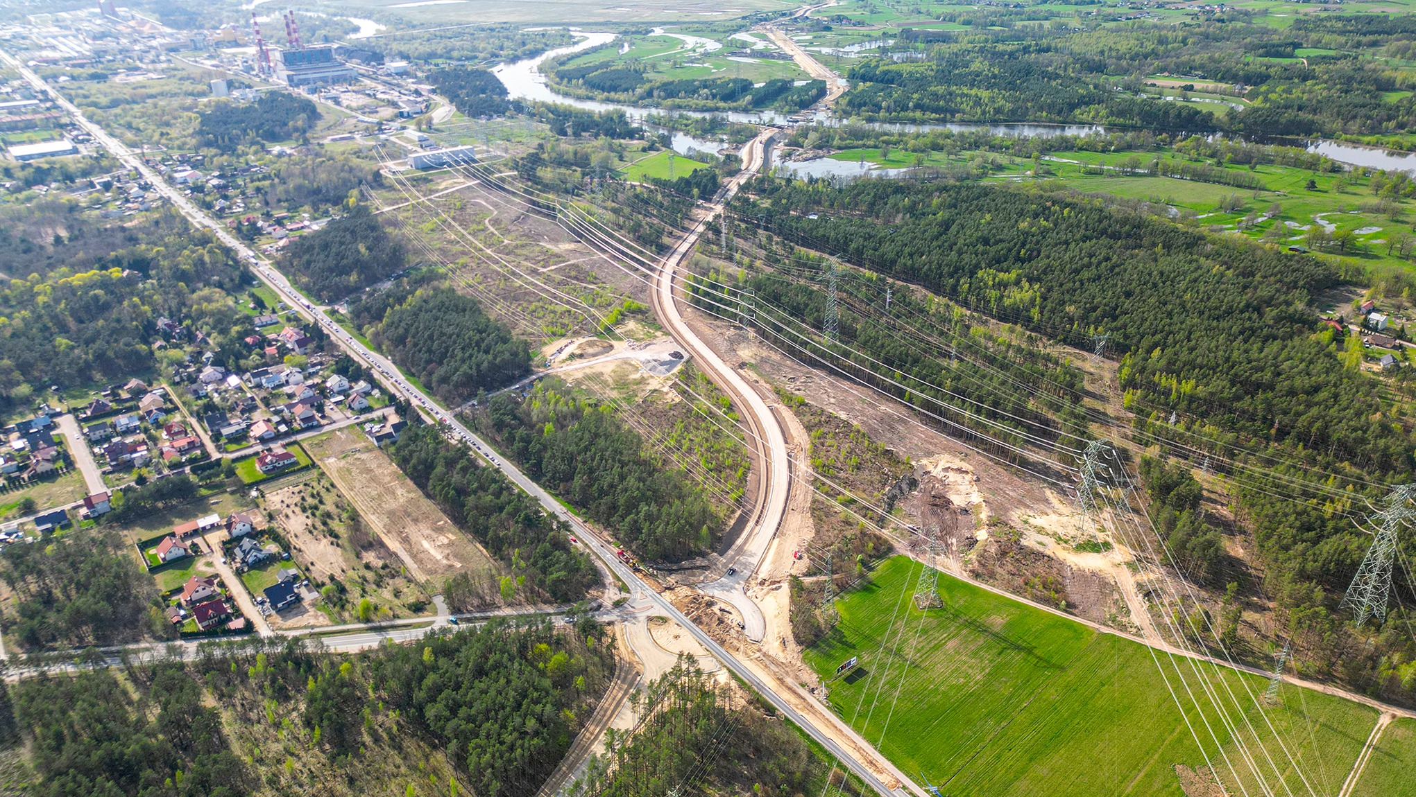 Zdjęcie z lotu ptaka przedstawia budowę nowej drogi ekspresowej przecinającej tereny zielone, lasy i rzekę, z widocznymi liniami wysokiego napięcia i fragmentami zabudowy mieszkalnej w tle.