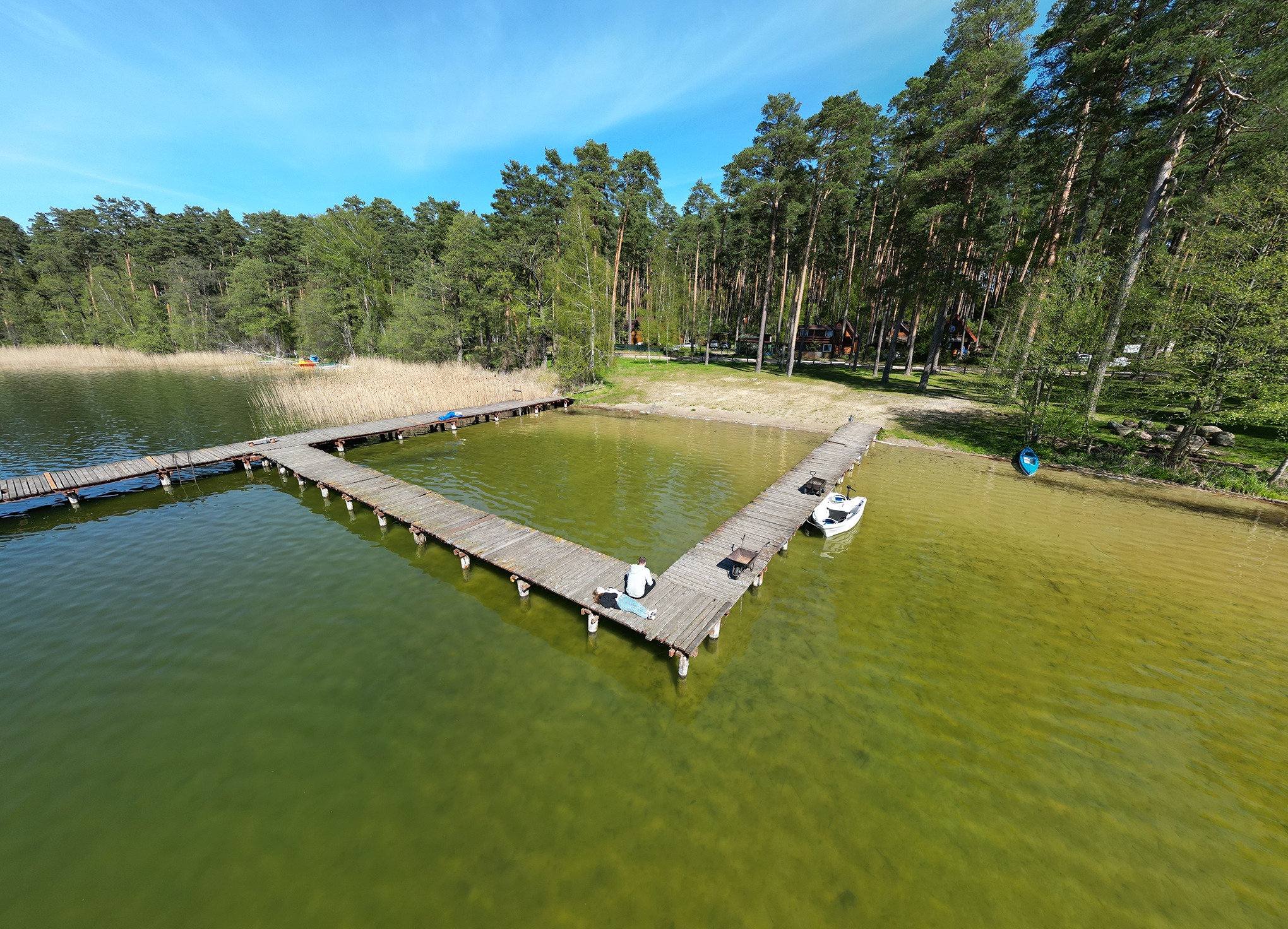 Widok z drona na drewniany pomost w kształcie litery L nad jeziorem w okolicach Ostrołęki, gdzie dwie osoby relaksują się na jego końcu, obok zaparkowana mała łódka.