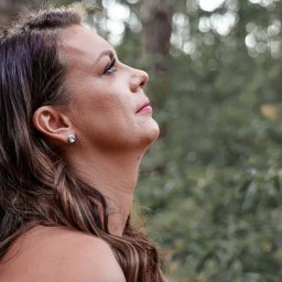 Krzysztof Raczyński - Fotografia - Portret kobiety opierającej się o korę drzewa, z widocznym kolczykiem z jasnym kamieniem i subtelnym makijażem, skierowanej wzrokiem ku górze na tle rozmytej zieleni.