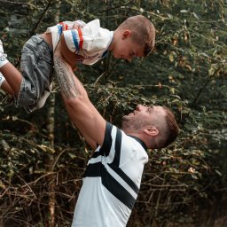 Krzysztof Raczyński - Fotografia - Tata podnosi synka do góry na tle zieleni, widoczne tatuaże na ramieniu ojca i zabawne skarpetki syna z motywem sowy.
