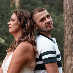 Krzysztof Raczyński - Fotografia - Para opiera się plecami o siebie na tle drzew: kobieta patrzy w lewo, mężczyzna spogląda w obiektyw.