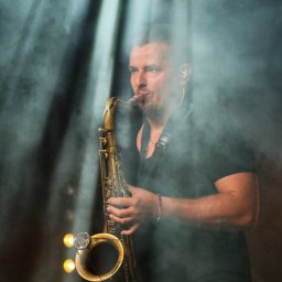 Krzysztof Raczyński - Fotografia - Saksofonista grający na saksofonie tenorowym, oświetlony reflektorami w zadymionym otoczeniu, widoczne słuchawki douszne