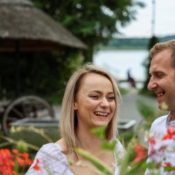 Krzysztof Raczyński - Fotografia - Uśmiechnięta para w plenerze, kobieta blondynka w białej sukience, mężczyzna w białej koszuli, w tle altana z trzciny i jezioro.