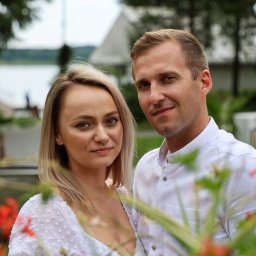 Krzysztof Raczyński - Fotografia - Portret pary w białych strojach na tle zieleni i czerwonych kwiatów, widoczny fragment jeziora i budynek w oddali.