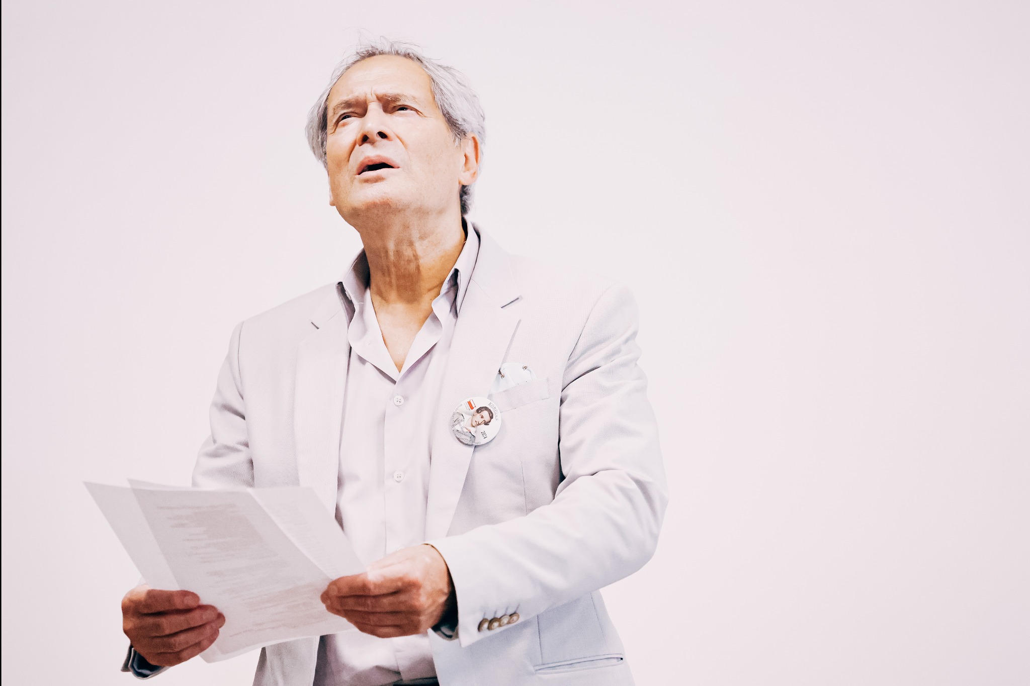 Jerzy Zelnik w jasnym garniturze, z przypinką Kordiana, czyta tekst z kartki, z głową uniesioną ku górze na tle jasnego tła.