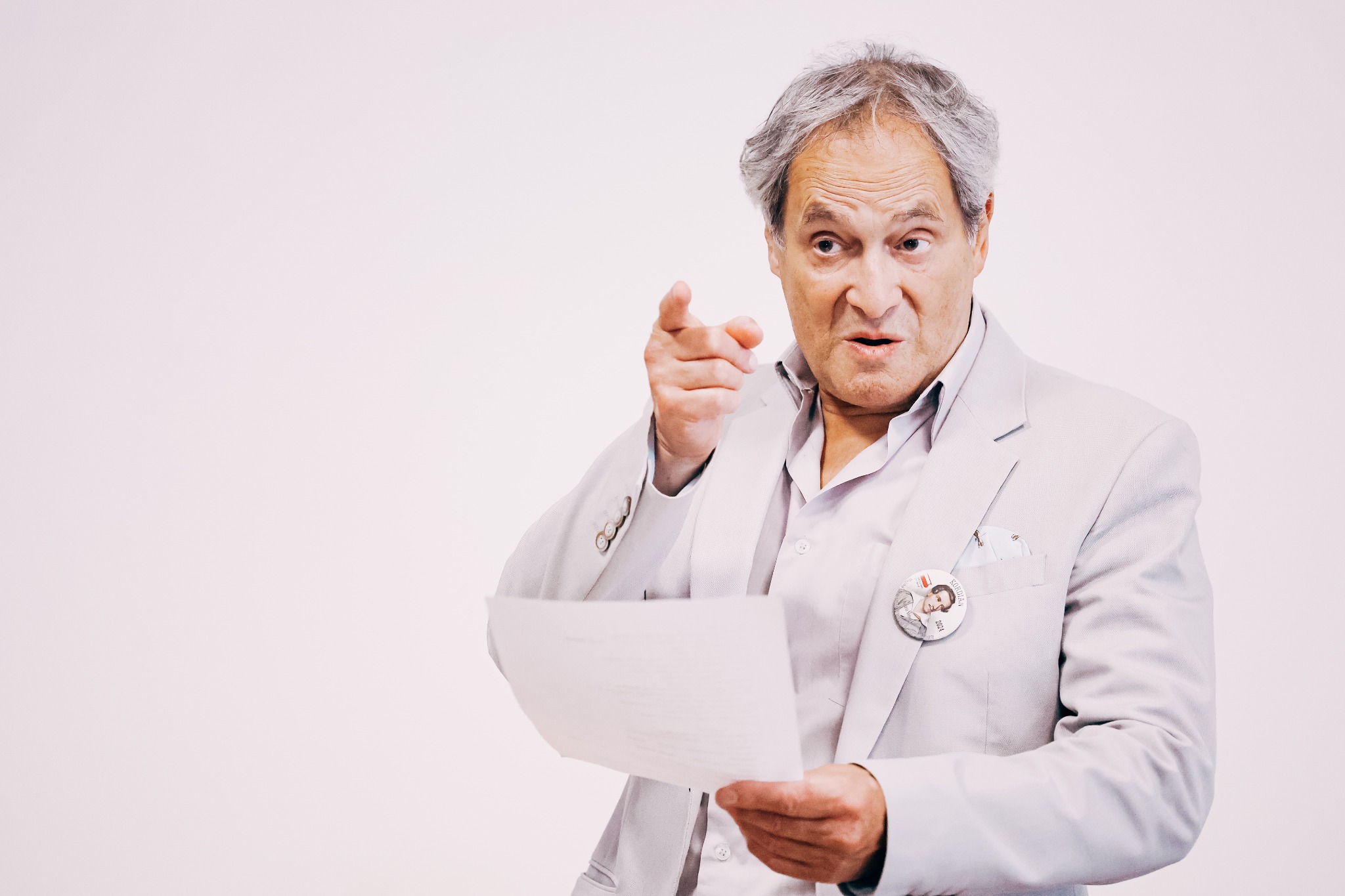 Aktor Jerzy Zelnik w jasnym garniturze, z przypinką 'Kordian 2024', czyta tekst z kartki i wskazuje palcem w stronę widza, na jasnym tle.