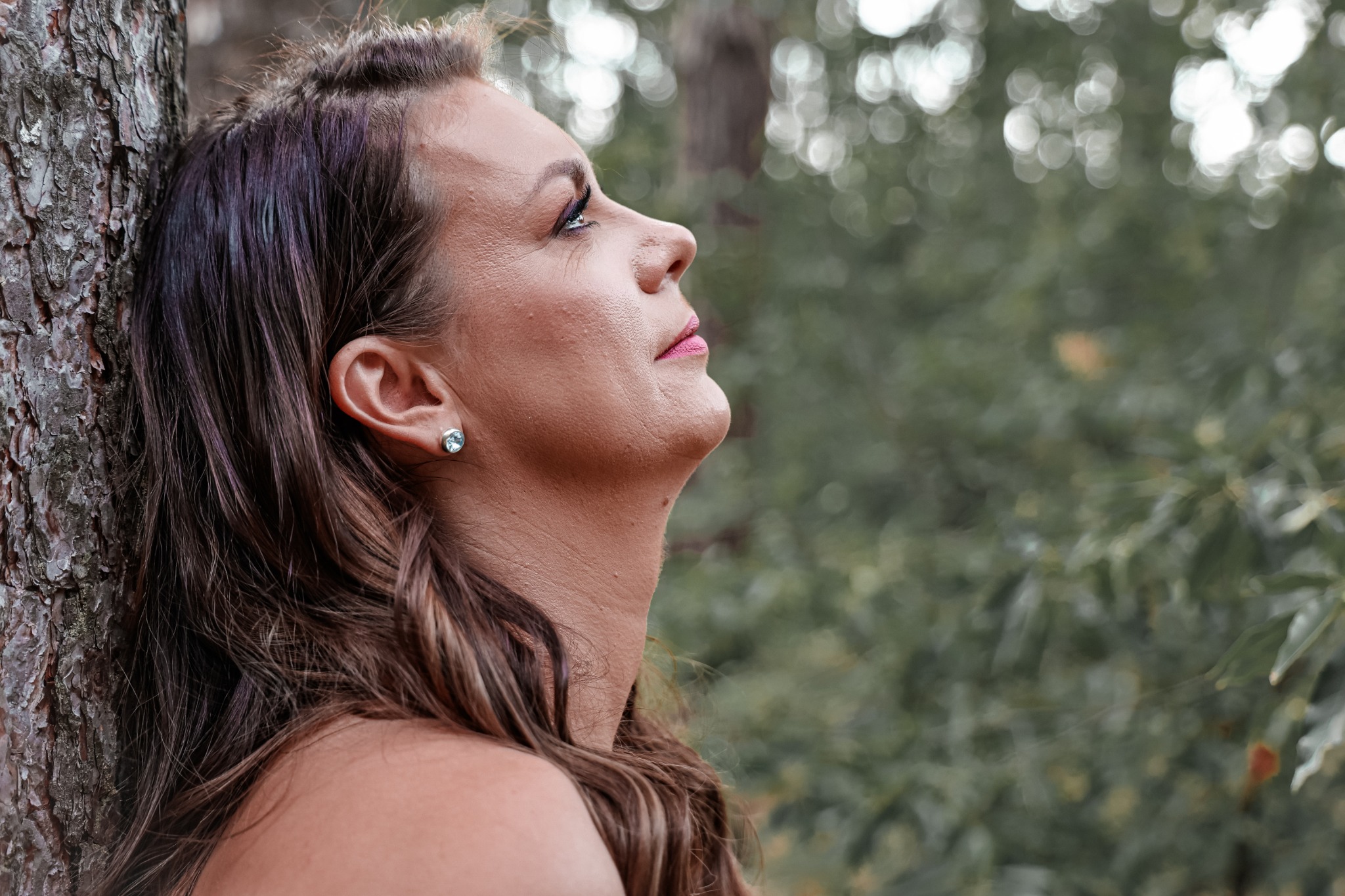 Portret kobiety opierającej się o korę drzewa, z widocznym kolczykiem z jasnym kamieniem i subtelnym makijażem, skierowanej wzrokiem ku górze na tle rozmytej zieleni.