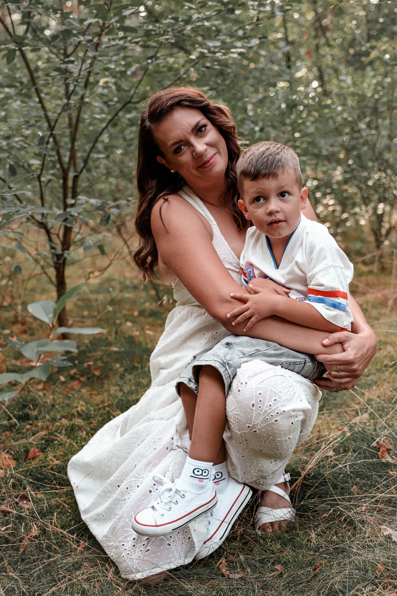 Matka w białej sukience trzyma synka na kolanach, oboje patrzą w górę, w tle las.